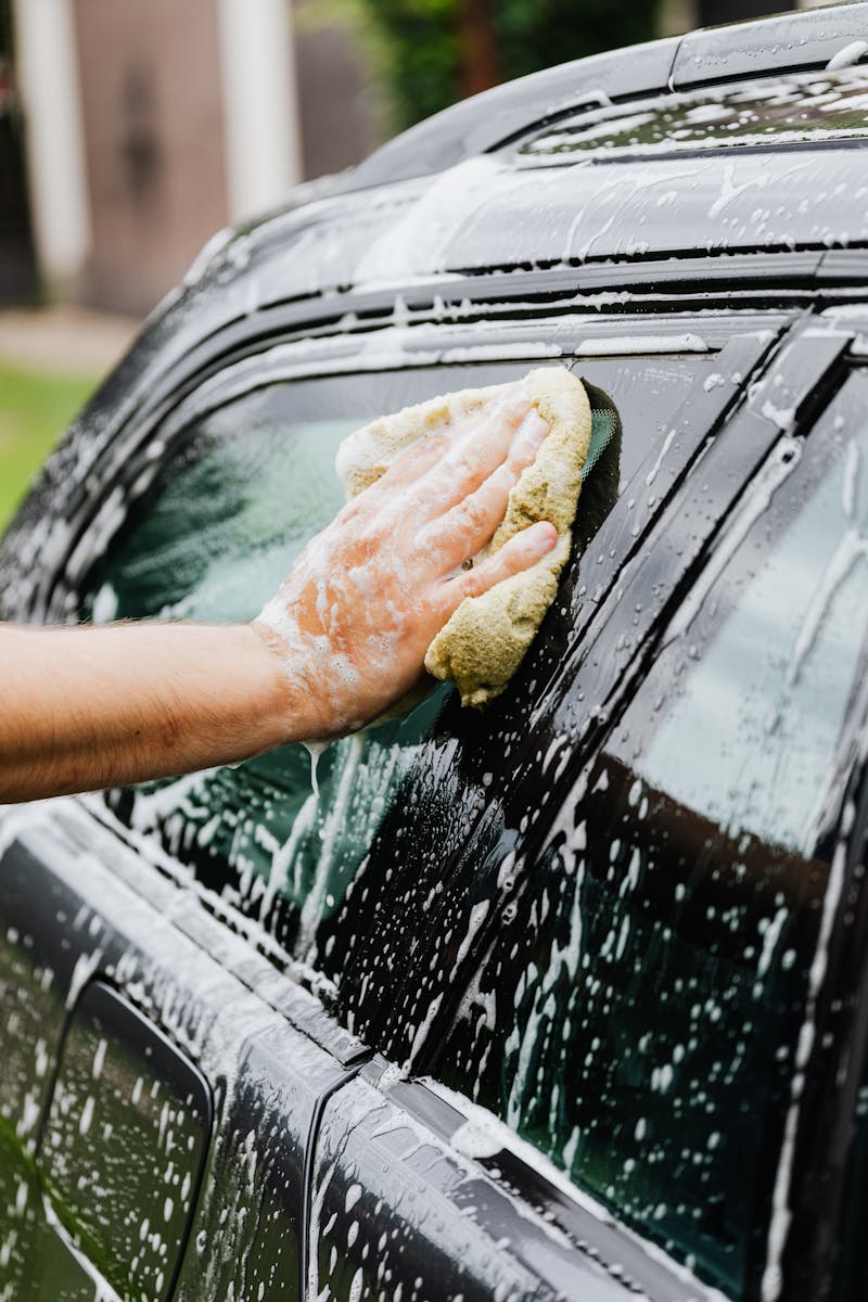Close-up of a person washing a car window with a sponge and soap suds outside.