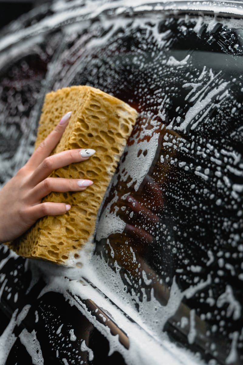 A hand scrubs a car window with a sponge, covered in soap and foam, at a car wash.
